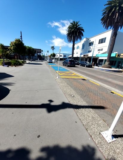 Wide, level concrete footpath alongside a level paved road. A section of the road is blue with yellow cross-hatching, indicating a restricted parking or no-standing zone.