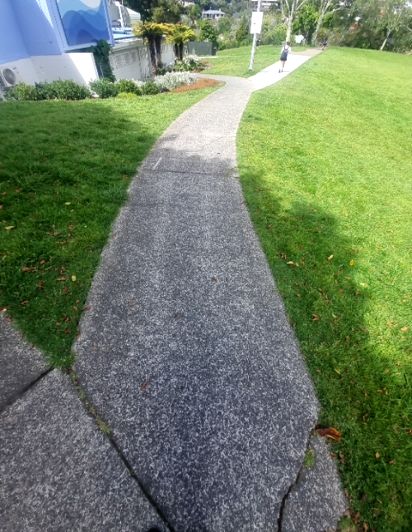 Speckled dark grey concrete pathway with a clear fork. Green grass verges border the level path. A distant person walks on an uphill section.
