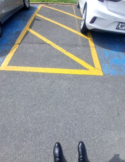 Asphalt car park with a yellow-lined, diagonally striped shared zone next to blue accessible parking markings. Partial views of two cars parked on either side.