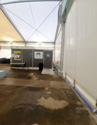 Wet, uneven concrete floor in a temporary tent. Ahead, a grey modular block features accessible family toilet signage and a small ramp to the entrance.