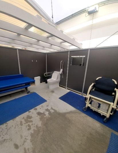 Accessible facility: toilet with grab rails, blue adjustable changing bench, and wheeled shower chair. Wet concrete floor with blue non-slip mats. Overhead light fixture.