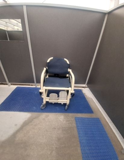 Shower cubicle with a wheeled commode chair on blue ribbed mats, wet concrete floor, a drainage channel, and a small mirror.