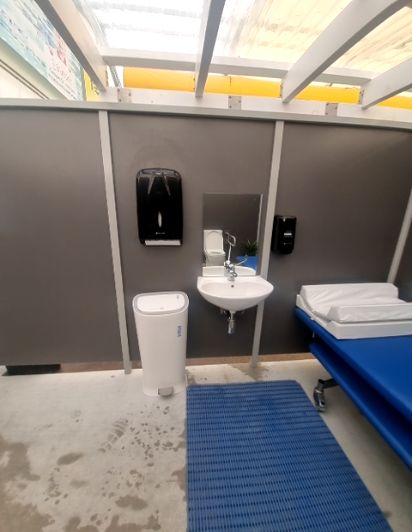 Wall-mounted sink, mirror, and dispensers in a grey-walled bay. White pedal bin and blue mobile changing bed with white mat. Wet concrete floor with footprints, partially covered by blue ribbed mat...