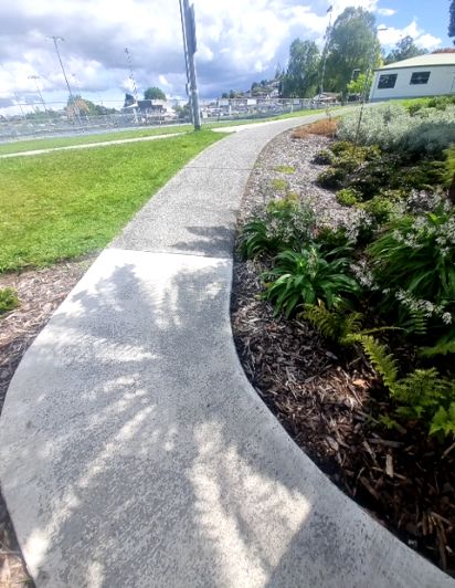 Curved, wide, grey textured concrete pathway with a gentle gradient, bordered by green grass on the left and a planted garden bed on the right. Tree shadows fall across the path.
