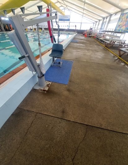 Indoor pool area with a grey accessible pool hoist on a blue mat, beside a cracked concrete pathway. Rows of wooden spectator seating are on the right.