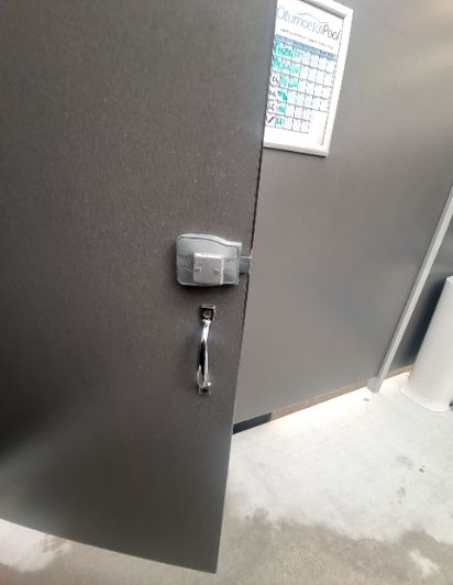 Dark grey partition door, slightly open, has a silver latch and a curved pull handle. The light concrete floor has wet patches, possibly a slip hazard.