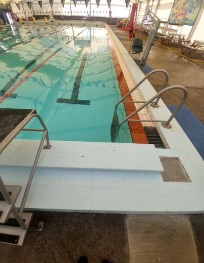 White tiled pool steps with stainless steel handrails lead into an indoor pool from a dark concrete deck. A pool hoist for assisted access is further right.