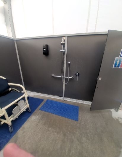 Roll-in shower with adjustable showerhead, two horizontal grab bars, and a wall-mounted soap dispenser. A shower chair rests on blue non-slip mats on a level concrete floor.