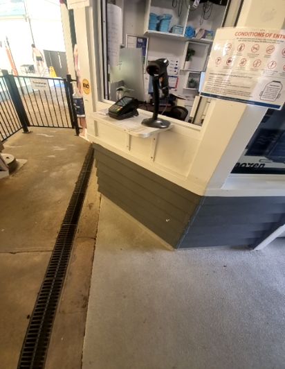 Level concrete pathway with a long grated drain. A standing-height counter features a payment terminal and "Conditions of Entry" signage. A black metal gate is on the left.