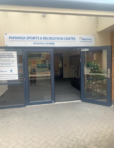 Papamoa Sports & Recreation Centre entrance with automatic glass doors, level herringbone-paved pathway, and informative signage.