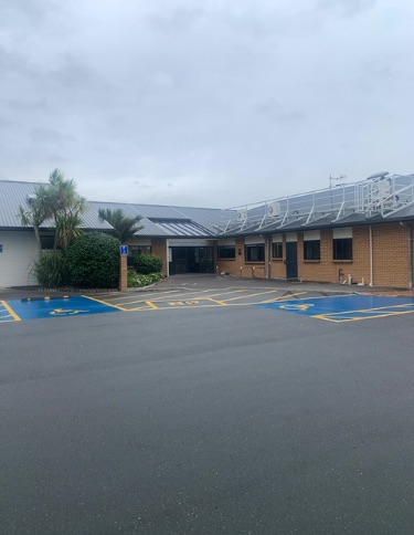 Asphalt carpark with three blue accessible parking bays, yellow markings. Level access to a building's main entrance, with a blue 'H' sign.