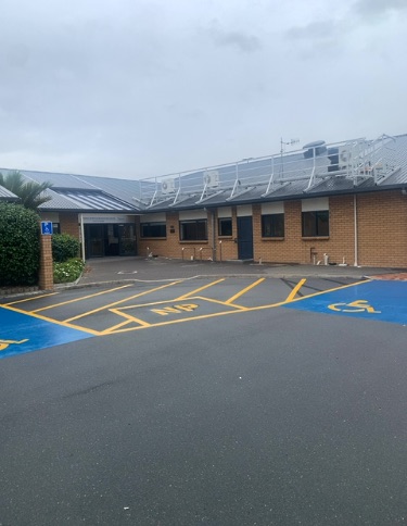 Asphalt car park features a blue accessible parking bay with a white wheelchair symbol, and a corresponding blue sign on a brick pillar. Leads to a brick building with a level, covered entrance.
