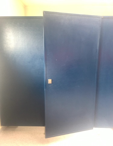 Dark blue toilet cubicle partitions. The centre door is ajar, revealing a small, light-coloured latch. Light-coloured floor surface.