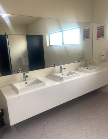 Washroom with three white washbasins on a floating white vanity. Chrome taps. Light speckled floor. Toilet cubicle reflected in large mirror. Bright window light.