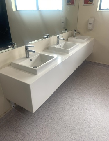 Three white sinks with chrome taps on a floating white vanity unit. Level light grey speckled floor. Two wall-mounted soap dispensers. Room is well-lit by natural light, potentially allowing wheelc...