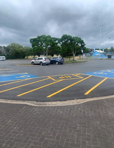 Accessible parking bay with blue markings and wheelchair symbol on asphalt, next to yellow diagonal lines. A paved footpath with a low kerb connects to the parking area.