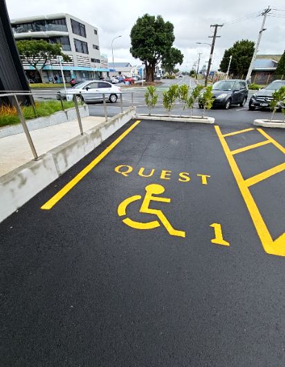 Accessible parking space 1, marked 'QUEST' on black asphalt with yellow lines. To the left is a concrete ramp with metal handrails, providing an accessible pathway.