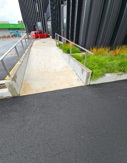 Concrete ramp with silver handrails on both sides, leading to a dark building entrance. Dark asphalt path connects to the ramp.
