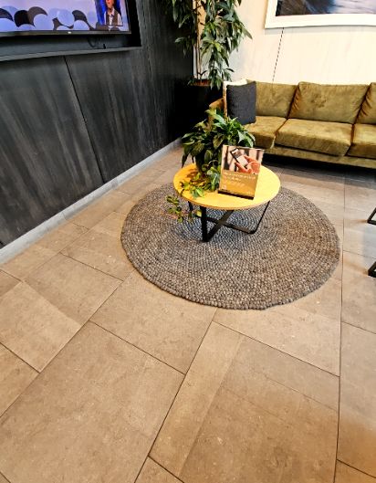 Level light brown tiled floor in a waiting area. An olive green sofa is against the wall. A round grey woven rug and low wooden coffee table on the floor form potential pathway obstructions.