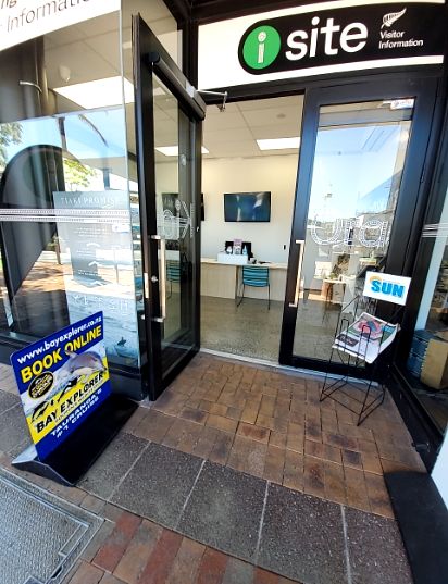 Level, multi-toned block paved footpath leads to an i-site visitor information centre with open double glass doors. An A-frame sign and newspaper rack are on the footpath.