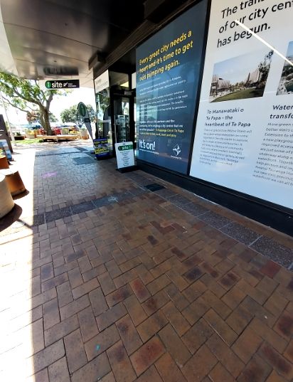 Wide, level footpath with herringbone brick pavers runs past a building. An A-frame 'i site' sign partially obstructs the path near a glass door entrance. Further along, outdoor benches are visible...