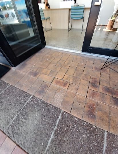 Glass double door entrance with a silver threshold. The outside pathway has uneven, raised terracotta pavers directly at the entry, followed by a darker aggregate path. Interior floor is level.