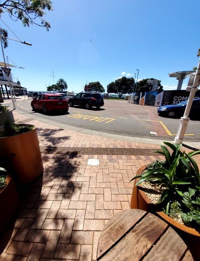 Level brick paved footpath with tree shadows and brown planters, bordering a curved road with a yellow 'LOADING' zone. In the distance are waterfront buildings and construction hoarding.