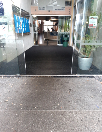 Level entrance from concrete pavement to a wide, dark grey ribbed floor mat. Automatic sliding glass doors lead to an indoor reception area with a level floor surface throughout.