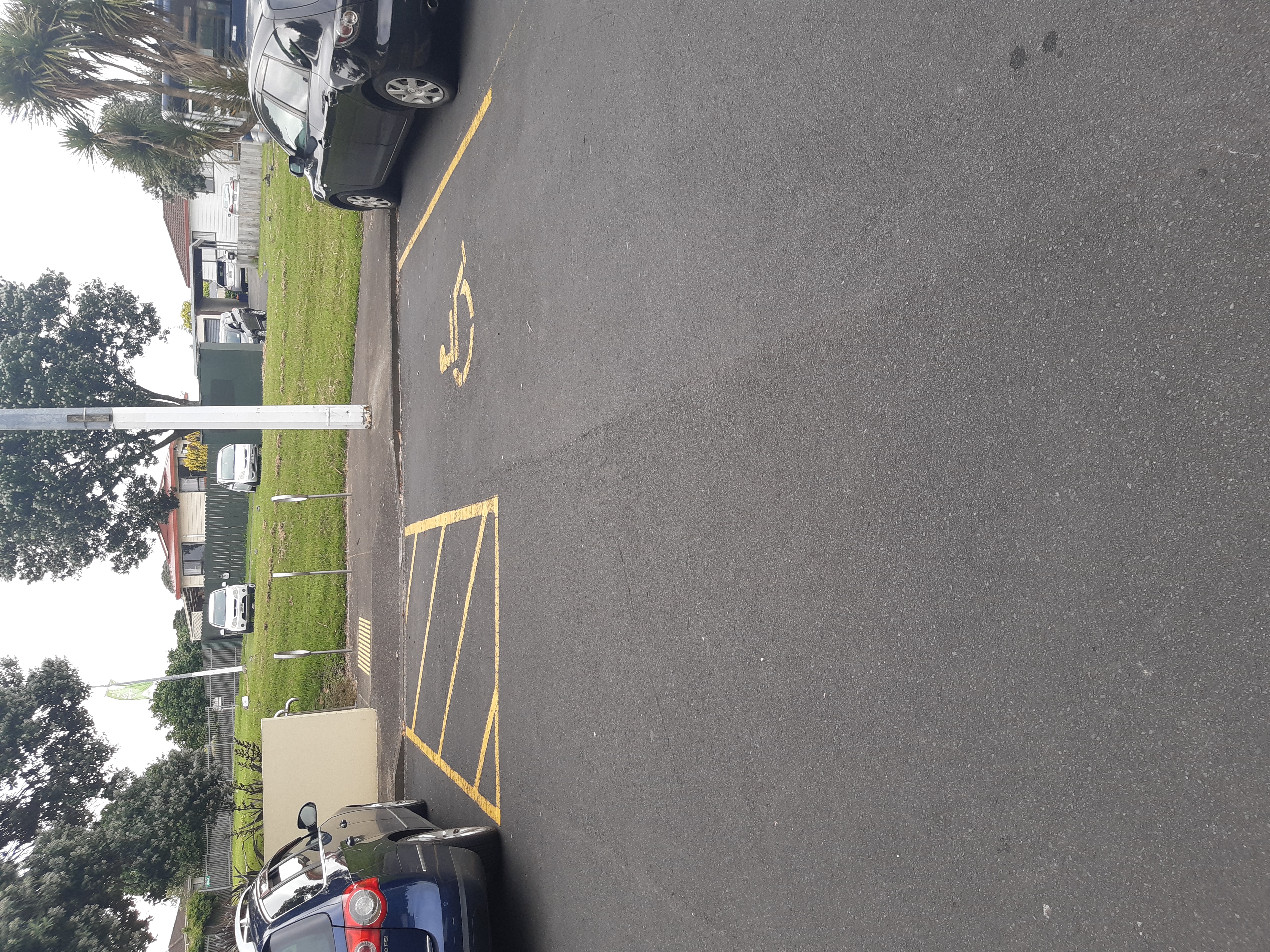 Accessible parking space with yellow wheelchair symbol and hatched shared zone on asphalt. Concrete kerb ramp with yellow tactile indicators leads to a level footpath.