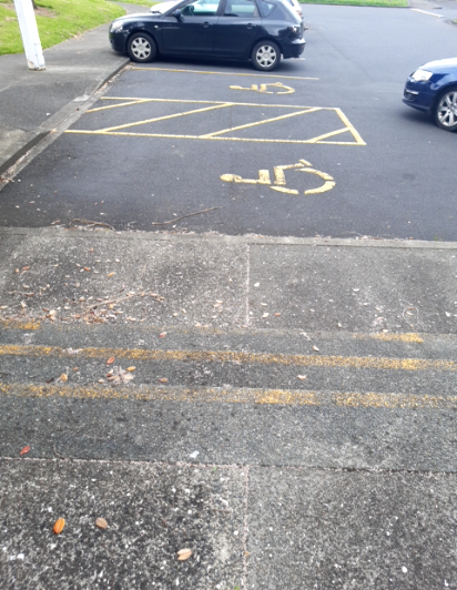 Two accessible parking spaces with yellow wheelchair symbols on level asphalt. A yellow hatched aisle borders the spaces, next to a weathered concrete path with yellow line markings.