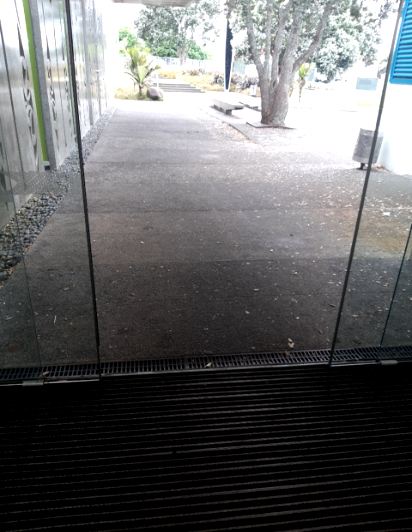 Level entrance through glass doors to a wide, flat concrete pathway. A black ribbed floor mat is inside, with a metal drainage grate at the threshold. Natural light throughout.
