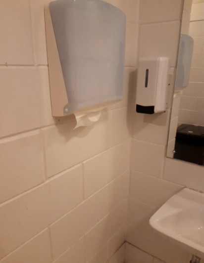 White tiled wall corner with a light blue paper towel dispenser, white soap dispenser mounted next to a mirror, and a small wall-mounted ceramic basin. No grab bars are visible.