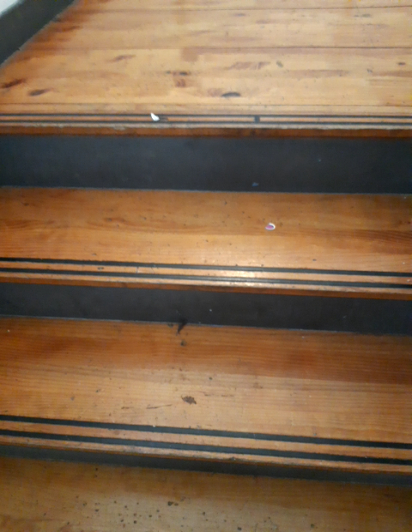 Polished wooden stairs with dark grey risers. The edge of each step features two dark parallel lines for visibility and grip. No handrails are visible.