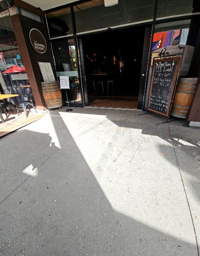 Level concrete pathway leads to a wide, step-free entrance with a slim metal threshold. Low wooden deck with outdoor seating to the left. Barrel and chalkboard sign stand to the right.