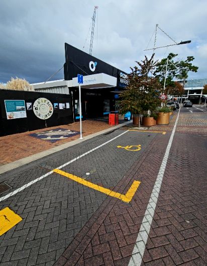 Accessible parking space with yellow wheelchair symbol on grey herringbone pavers. Level brick footpath with blue and white sign on white pole. Wide, level path leading to building entrance.