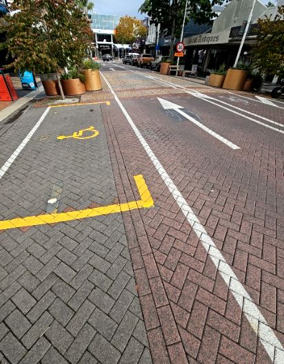 Level grey herringbone accessible parking with a yellow wheelchair symbol. A white line separates it from a red-paved roadway. Shopfronts and planter boxes line the level pedestrian areas.