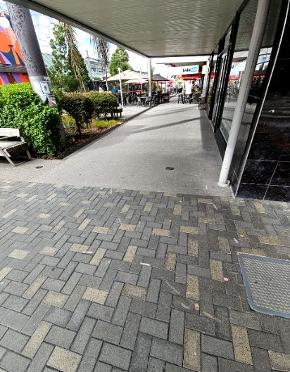 Wide, level pathway transitions from grey herringbone pavers to smooth concrete under a flat awning. A wooden bench is on the left. Clear access leads to outdoor cafe seating under umbrellas.