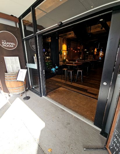 Wide, level entrance with a black-framed sliding door. Smooth concrete pathway leads to a flush floor mat and dark timber flooring inside. High-top tables and stools are visible within.