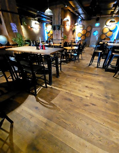 Unobstructed level timber floor. High wooden tables with tall black stools and some standard-height tables. Dimly lit by warm overhead pendant lights.