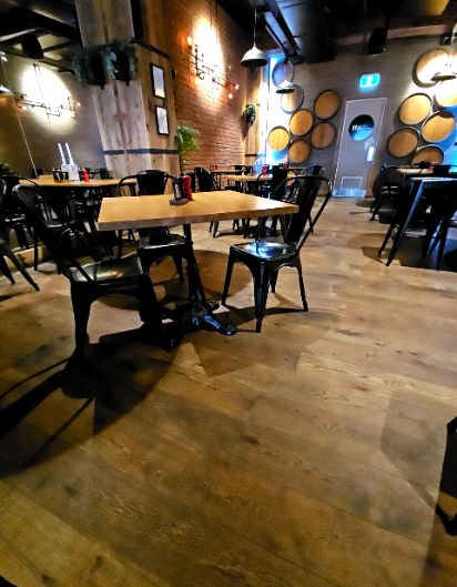 Level light timber floor with wide pathways. Black metal chairs and wooden tables are spaced throughout. Warm lighting from wall sconces and pendants. A clear route leads to a rear doorway.