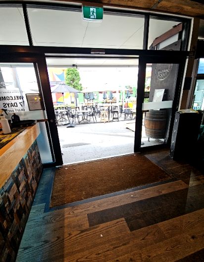 Level entrance through wide glass sliding doors with a flat coir mat. Timber-look flooring leads to a level outdoor patio with tables and chairs. Green exit sign mounted above the doorway.