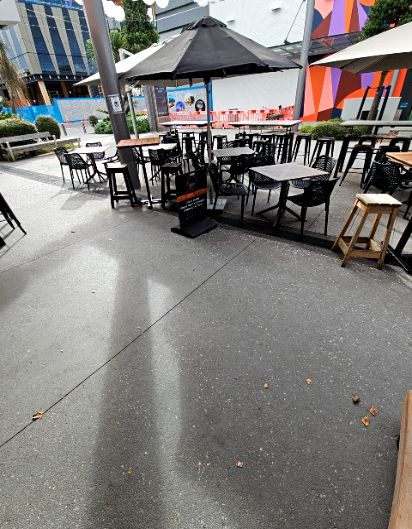 Level grey concrete pavement with wide pathways between outdoor cafe seating. Black metal tables, chairs, wooden stools, and large black umbrellas provide shade. Small black A-frame sign.