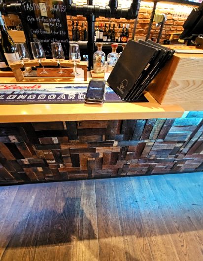 Level light timber floor meets a standard height light timber bar counter. The bar front is a textured wood block mosaic with blue under-counter lighting. No lowered section or seating is visible.
