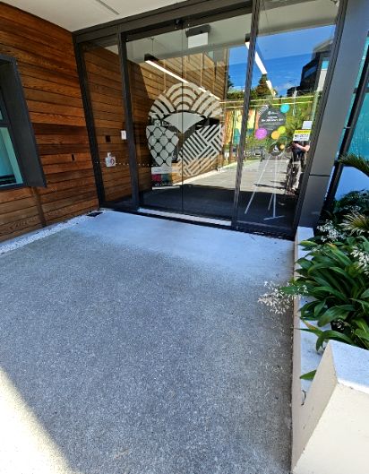 Wide, level concrete pathway approaches automatic glass doors with a decorative pattern. The entrance is bordered by a timber wall and a low planter box.
