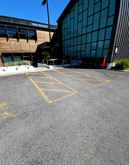 Asphalt foreground with yellow hatched zones. A sloped pathway, edged by a white planter, leads to an entrance between two modern buildings. An orange cone is near the right building.
