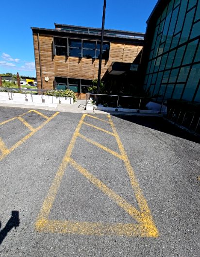 Asphalt car park with yellow hatched markings. Concrete pathway with metal handrails leads to a building entrance between timber and glass-fronted sections.
