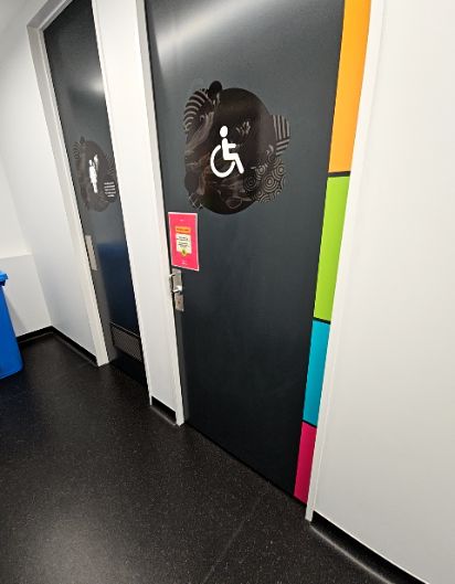 Dark accessible toilet door with a white wheelchair symbol and colourful side trim. Next to a standard toilet door. Level dark speckled floor pathway.