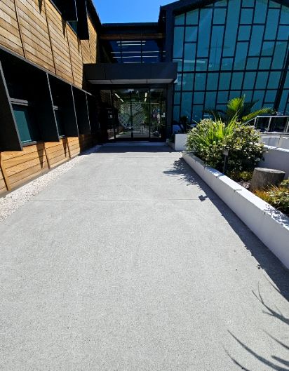 Wide, level concrete pathway leading to automatic glass doors of a modern building. Wood panelled facade on left, reflective glass facade on right. Landscaped area with low white planter box next t...