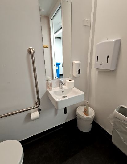 Accessible toilet cubicle with a long, silver grab bar on the left wall and a wall-mounted white sink. Dark floor and white walls.