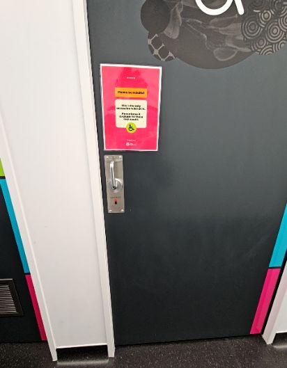 Dark grey accessible toilet door with a wheelchair symbol sign, silver entry button, and a level dark floor.